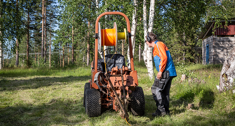 En orange maskin står ute på en äng. Den gräver i marken. En man står bredvid och tittar när den arbetar.