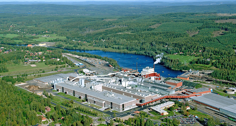 En stor fabrik ligger mitt i ett grönt skogslandskap, intill ett blått vatten.