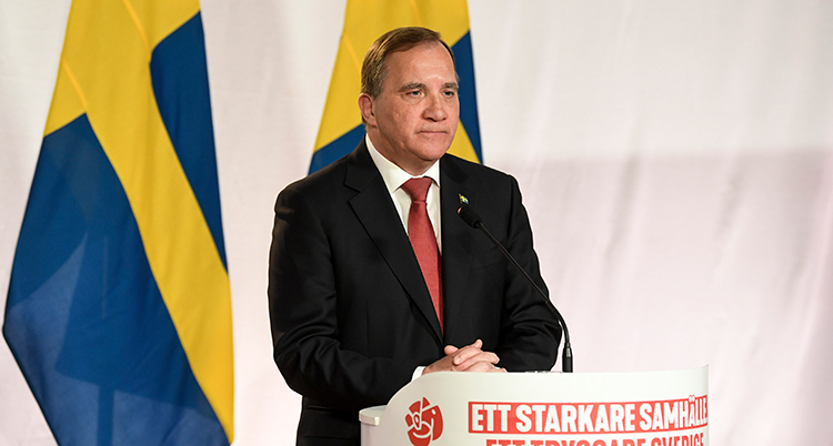Han står i en talarstol dekorerad med socialdemokraternas ros, har mörk kostym och röd slips, bakom syns svenska flaggor