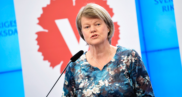 Hon har en mönstrad tröja och pratar i en mikrofon. Bakom syns Vänsterpartiets logga.