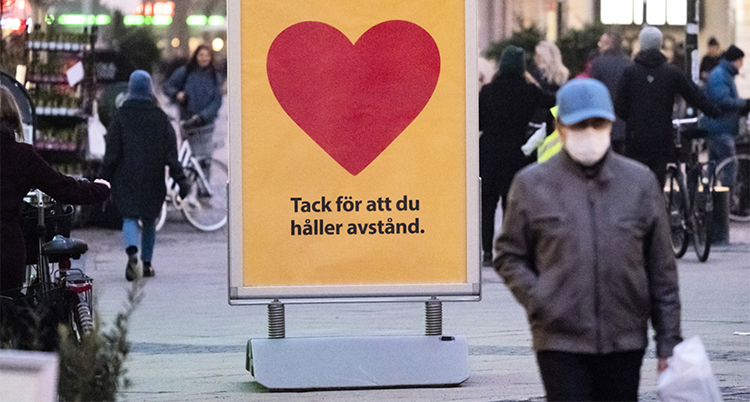 En stor skylt som säger: Tack för att du håller avstånd. En människa med munskydd går bredvid på gatan.