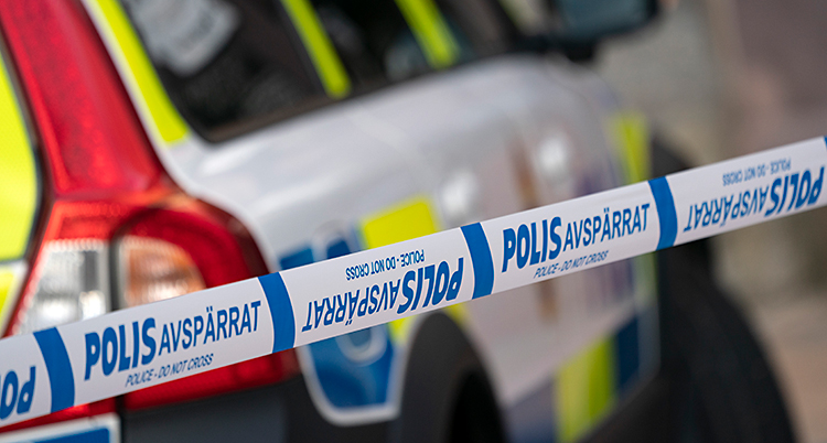 Poliser har spärrat av en plats. De har spärrat av med ett band som är blått och vitt. Bakom bandet står en polisbil.