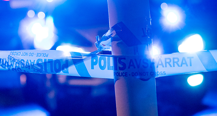 Det är utomhus. Det är mörkt. Poliser har satt upp avspärrningsband runt en lyktstolpe. I bakgrunden syns blåljus från en polisbil.