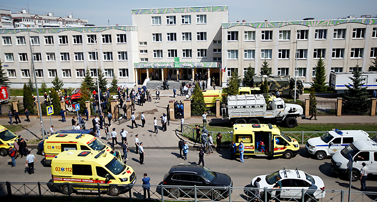 Bilden är tagen utanför en stor skola. Skolan är grå. På vägen utanför skolan finns flera ambulanser och polisbilar. Många människor är också där.