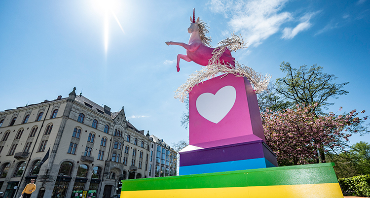 Statyn av en rosa enhörning står på en rosa- och regnbågsfärgad sockel. Den ses mot en blå himmel, träd icg hus i bakgrunden.