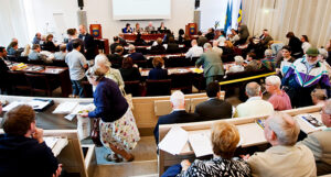 En sal som är full med folk. De sitter i bänkar. Längst fram sitter fyra personer på en egen bänk. De leder mötet.