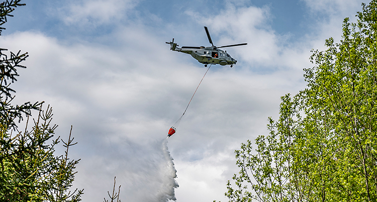 En helikopter flyger i luften. Från helikoptern hänger en lina. I slutet av linan finns en behållare som släpper ner vatten på marken.