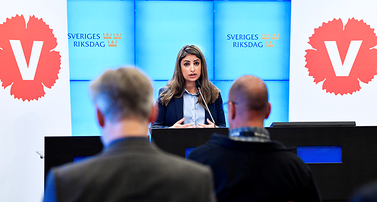 Nooshi Dadgostar står och pratar för journalister. Hon är i ett rum i riksdagen. Bredvid henne finns Vänsterpartiets symbol, en röd blomma.