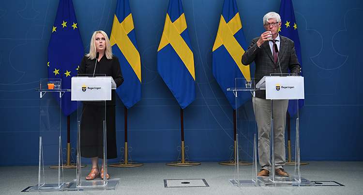 De står i varsin talarstol. Lena Hallengren står till vänster. Johan Carlson står till höger. Bakom dem finns svenska flaggor.