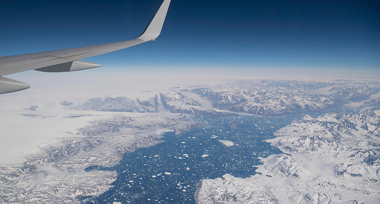 Vit is som ses från luften. Blå himmel och blått hav. En del av en flygplansvinge syns till vänster i bild