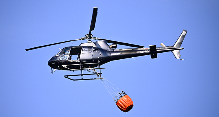 En helikopter i luften. Under den hänger en stor röd säck.