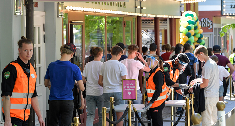Människor vid en ingång. de är på vägin och personer i orange västar kontrollerar biljetter. En av dem har visir.