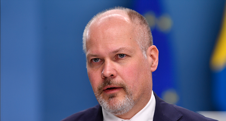 En nära bild på ministern. Han är skallig och har lite grått skägg.Bakgrunden är blå.