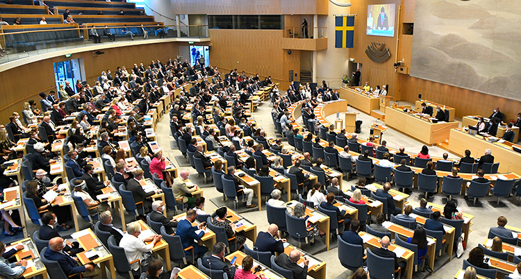 En bild över hela riksdagen sal. Politikerna sitter två och två i sina bänkar.
