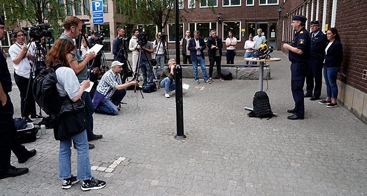 Tre poliser står utanför ett hus. De har ett litet bord där det står mikrofoner. Framför dem finns en massa journalister med anteckningsblock och kameror.