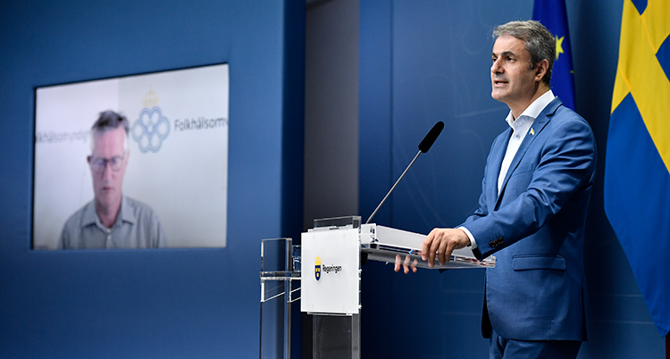 Ibrahim Baylan från regeringen pratar på presskonferens. Bredvid honom en tv-skärm där Anders Tegnell syns.