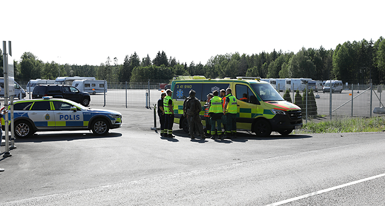 En polisbil och en ambulans står vid en väg. Flera personer står och pratar. Det finns stängsel och staket i bakgrunden.