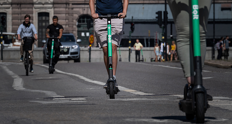 En bild från en gata i Stockholm där riksdagen syns i bakgrunden. I förgrunden syns underdelen av två gröna elsparkcyklar. En bit bort kommer två personer på cyklar.