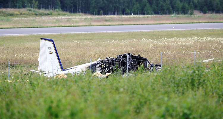 Flygplanet ligger på marken i gräset. Fotot är taget en bit ifrån. Fören på flygplanet är utbrunnet. Bakdelen ser intakt ut.