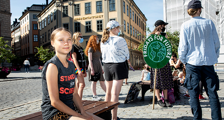 Hon har shorts och t-shirt och sitter på en bänkoch tittar snett in mot kameran. Bakom syns fler unga som står på ett torg i staden.