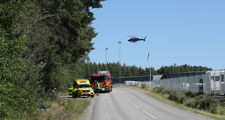 En gul ambulans och en röd bil från räddningstjänsten står vid sidan av vägen, en helikopter flyger i luften och på höger sida ses en del av det grå fängelset bakom ett nätstängsel.