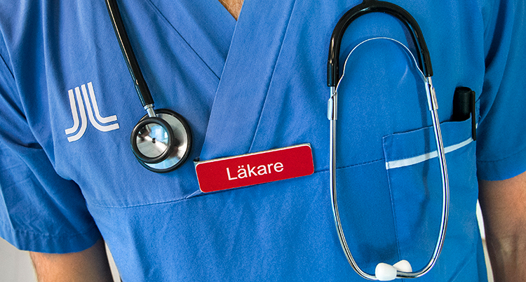 ´Läkare`står det på en röd namnskylt som sitter på en blå tunika. Runt halsen har personen ett stetoskop. Bara en del av överkroppen syns.