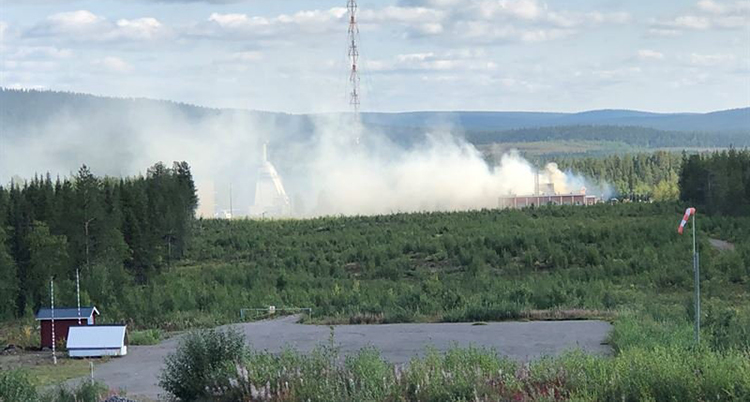 Rök syns över skog i en fjällmiljö. I förgrunden syns en asfalterad platta och en röd bod.