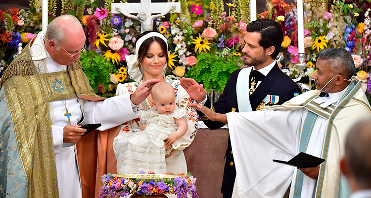 Julian i dopklänning blir döpt. Två präster, Sofia och Carl Philip står runt om honom. Han är i Sofias famn. Bakom dem är en fond av färgglada vackra blommor.