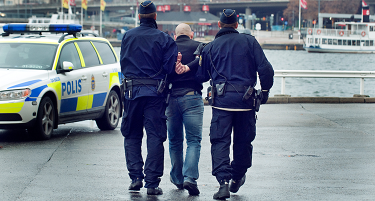 Två poliser håller fast en man och går mot en polisbil.