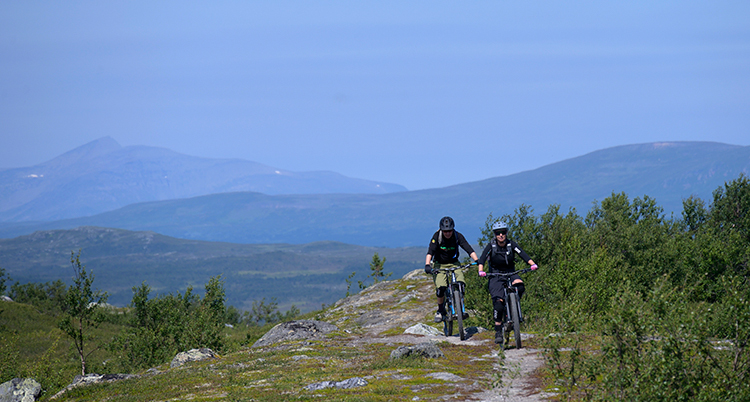 Två cyklister på en stig i fjällen, med fjäll i bakgrunden
