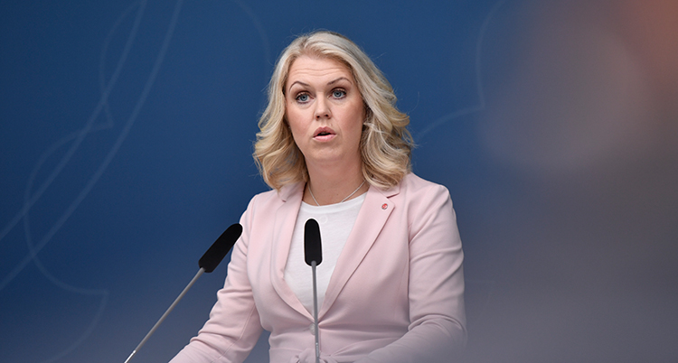 Hon står framför en mikrofon. Hon har ljusrosa kavaj.