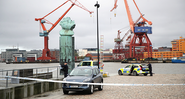 Stora lyftkranar syns i bakgrunden. Polistejp och polisbilar syns på piren.