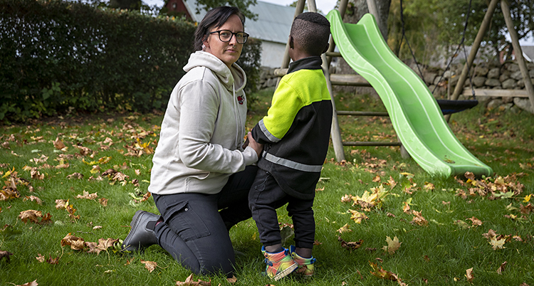 En kvinna och en liten pojke är på en gräsmatta. Kvinnan tittar in i kameran. Pojken tittar bort.