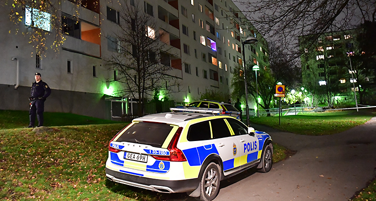 Det är mörkt. En polisbil och en polis står utanför ett stort hus med lägenheter.
