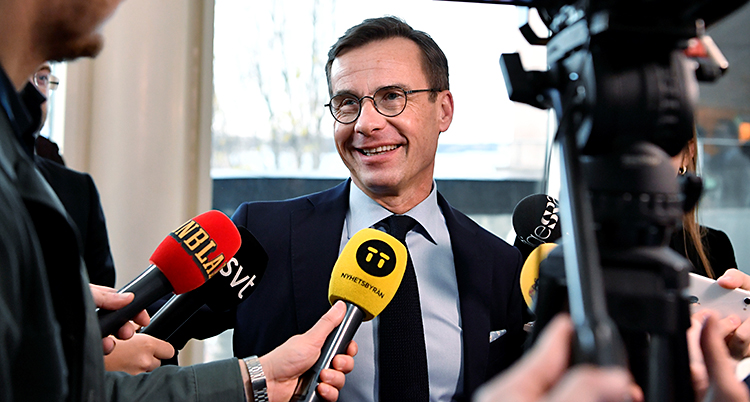 Ulf Kristersson pratar med journalister och ser glad ut. Han har en mörk kostym.