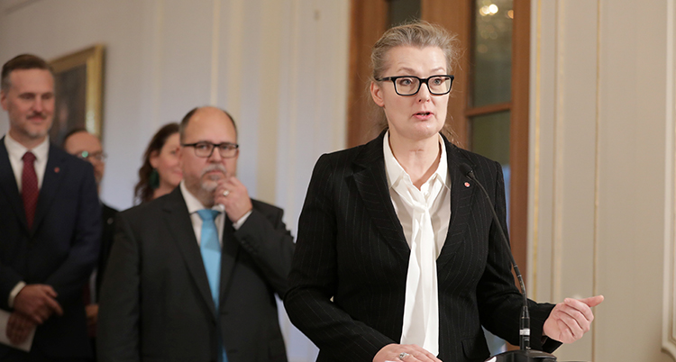 Hon står och talar. Hon har glasögon och svart kavaj.