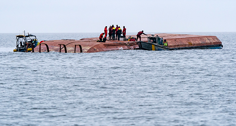 Ett fartyg ligger och flyter på havet. Fartyget ligger upp och ner. Flera personer står på fartyget.