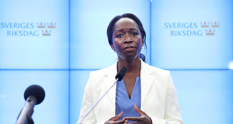 Hon står bredvid en mikrofon. Hon bär vit kavaj.
