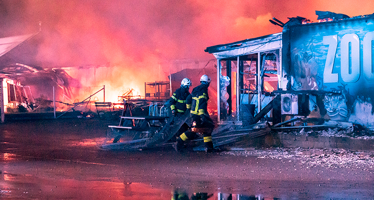 Rosaröda eldslågor syns mot himlen runt en trasig byggnad. Två räddningsklädda personer med hjälm går mitt i bilden.