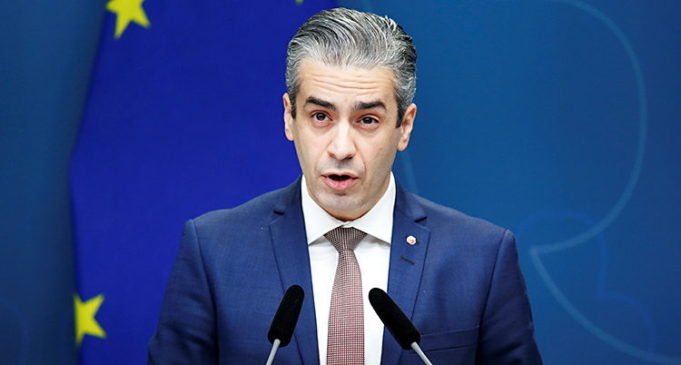 Han har kostym, slips och vit skjorta. Han tittar in i kameran och pratar i en mikrofon.
