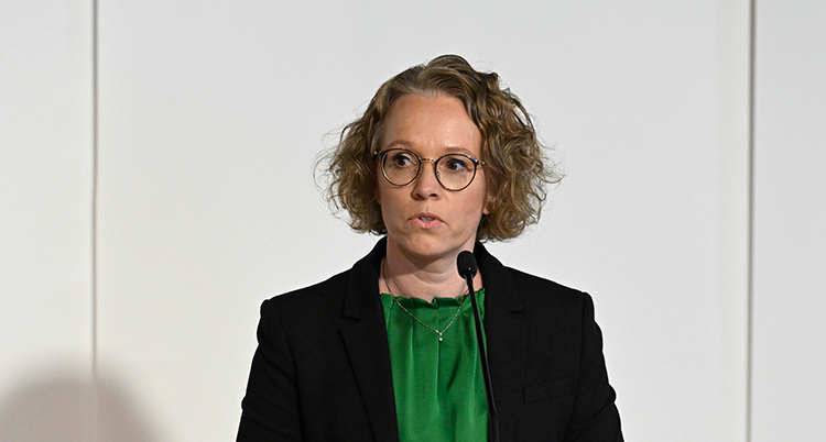 Hon pratar i en liten mikrofon. Hon har ljust, lockigt hår, glasögon, grön blus och svart kavaj.