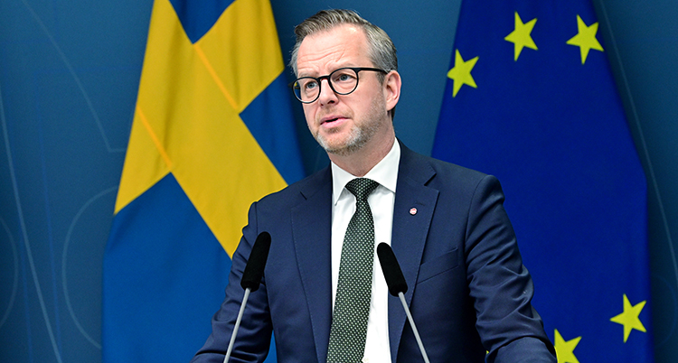 Han pratar i mikrofoner. Han har kostym, slips och glasögon.