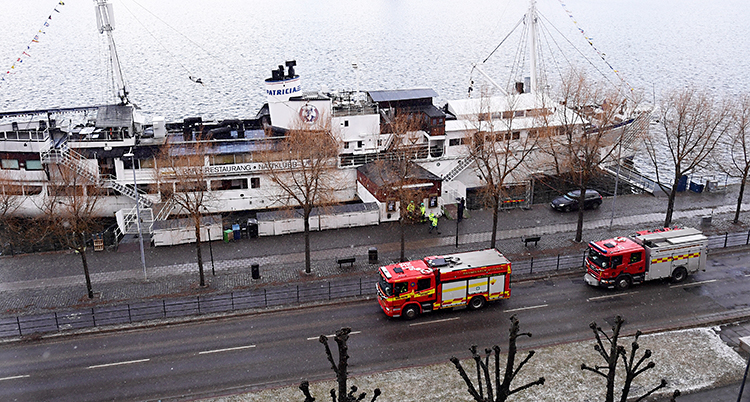 Bilden är tagen uppifrån en höjd, ner mot en väg och ett vatten. På vägen står brandbilar och vid kajen ligger en stor båt.