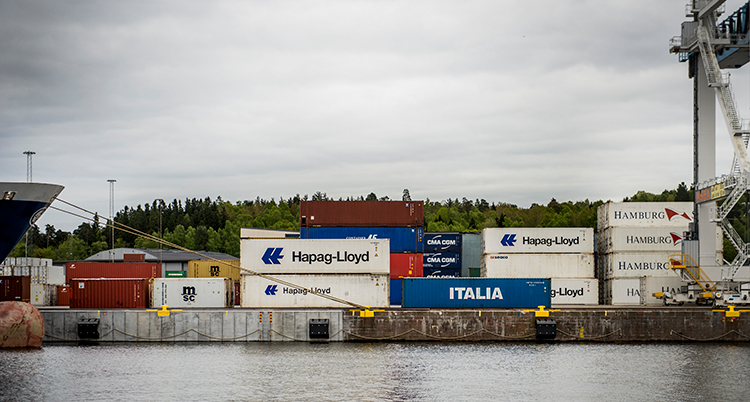 Containrar ligger lastad spå varandra på kajen. En del av ett stort fartyg syns till vänder. En travers syns till höger.