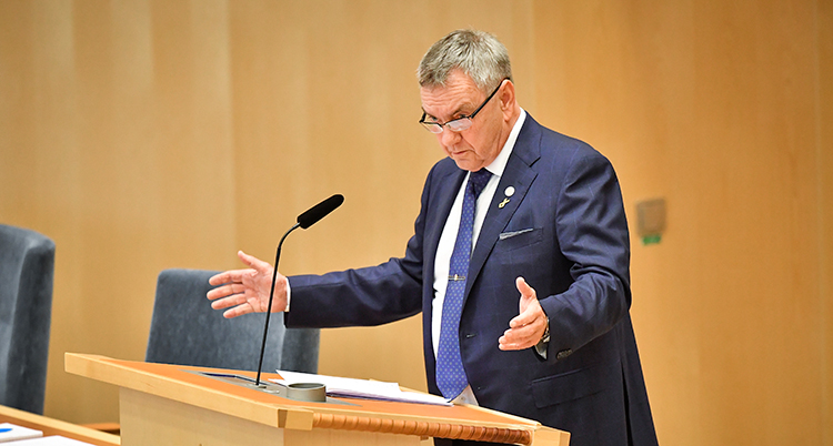 Han står i riksdagens talarstol och pratar. Han gör gester med båda händerna.