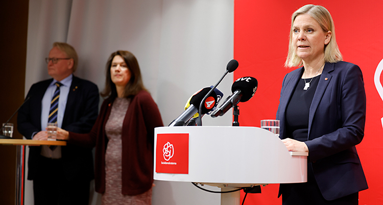 Magdalena Andersson pratar i mikrofoner på en träff med journalister. En bit bort står Peter Hultqvist och Ann Linde.
