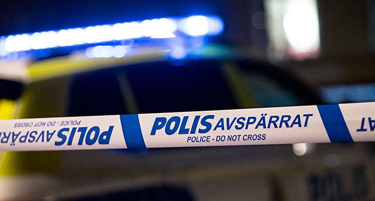 Det är mörkt ute. Poliser har spärrat av med ett band som är vitt och blått. I bakgrunden syns en polisbil.