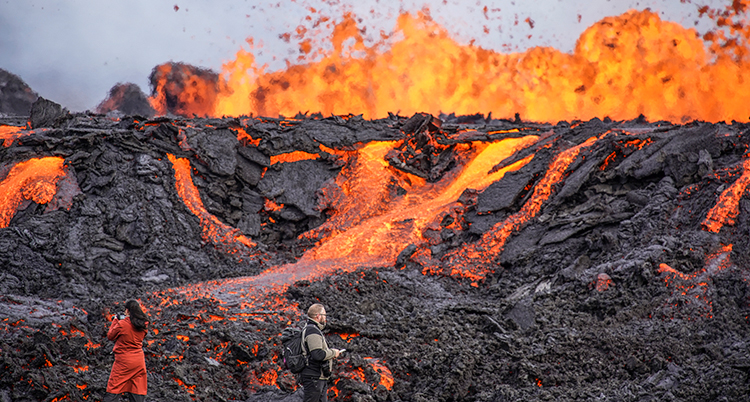 Röd lava rinner på den svarta fjällsidan och eldsflammor syns i bakgrunden. Längst fram står två personer och tittar.