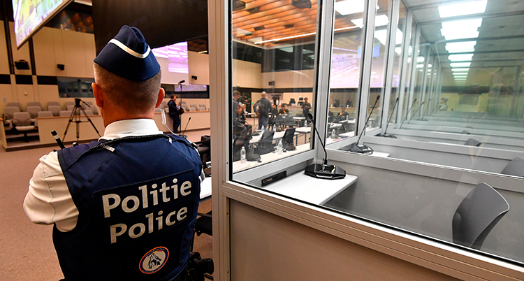 En polis står med ryggen mot kameran och ansiktet mot rättegångssalen. Han lutar sig mot ett bås med glasfönster där de åtalade ska sitta.
