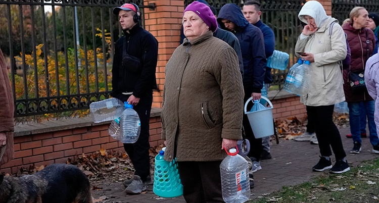 Människor står på en trottoar och väntar. De har ytterkläder. De håller olika platsbehållare i händerna.